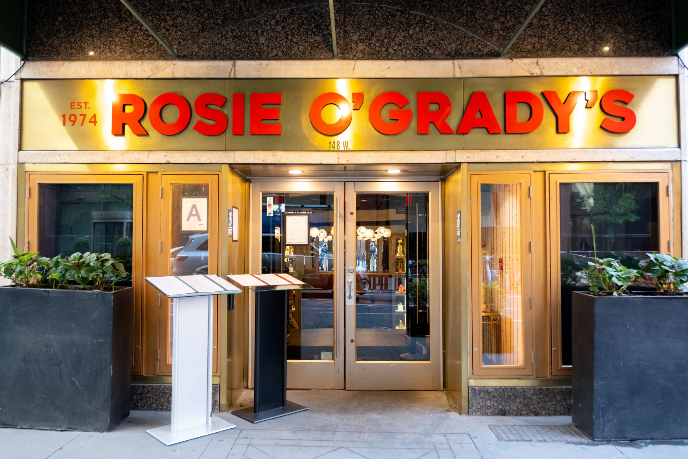 The illuminated Rosie O'Grady's Est. 1974 sign over a golden entrance with planters and menu stands.