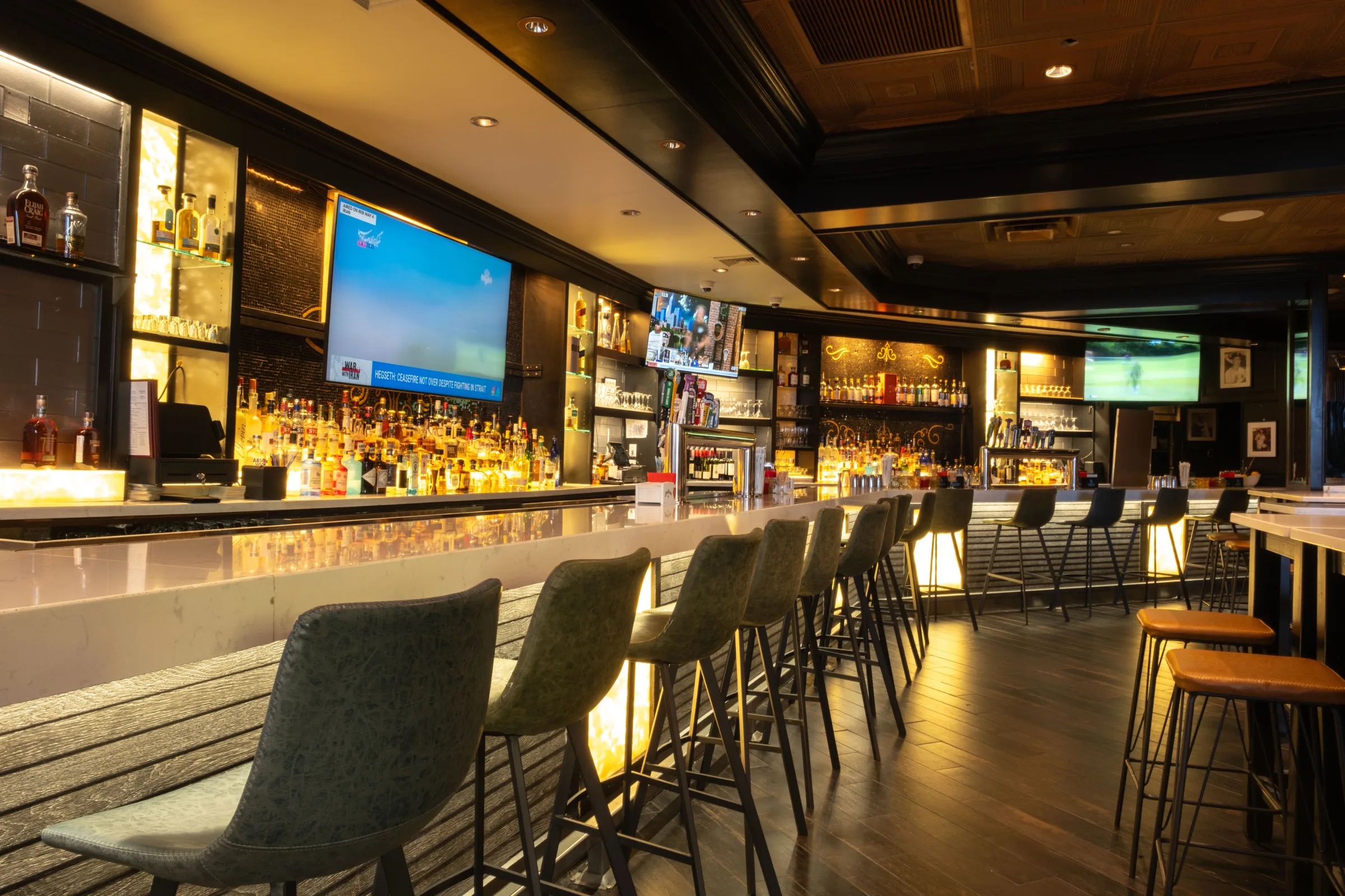 A long marble bar with tall stools, back-lit bottle shelves and TVs under a coffered brass ceiling.