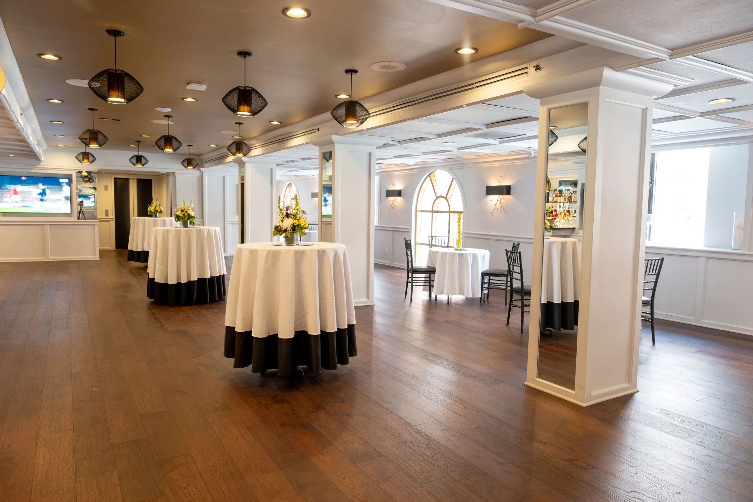 A wide view of an event room with a gold ceiling, white columns, arched windows and cocktail tables on a dark wood floor.