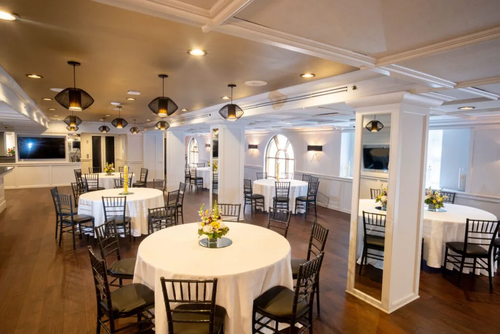 Round tables with flowers and black chairs across a dark wood floor, with mirrored white columns and arched windows.