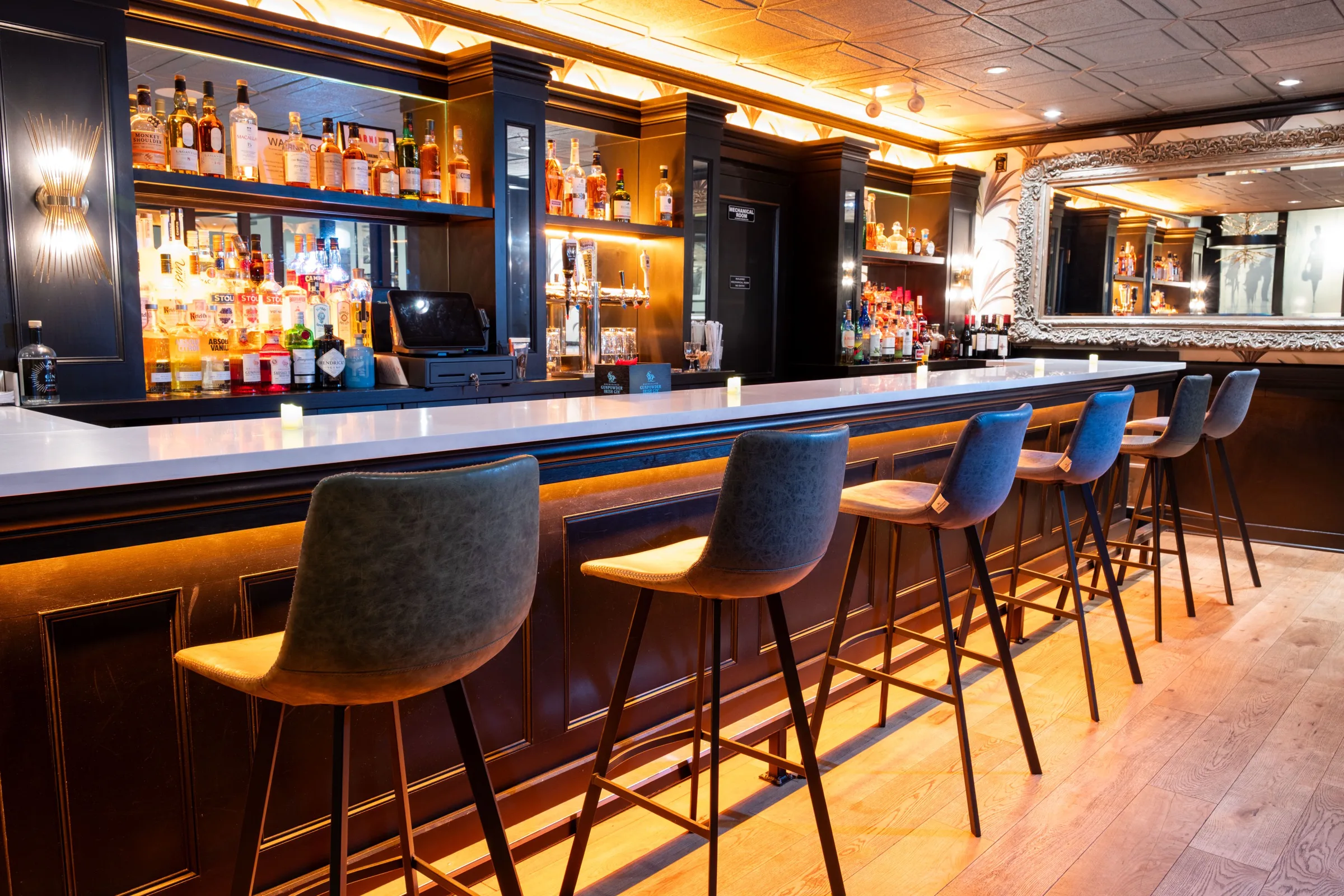 A bar with gray stools, warm under-bar lighting, bottle shelves and a large ornate mirror.