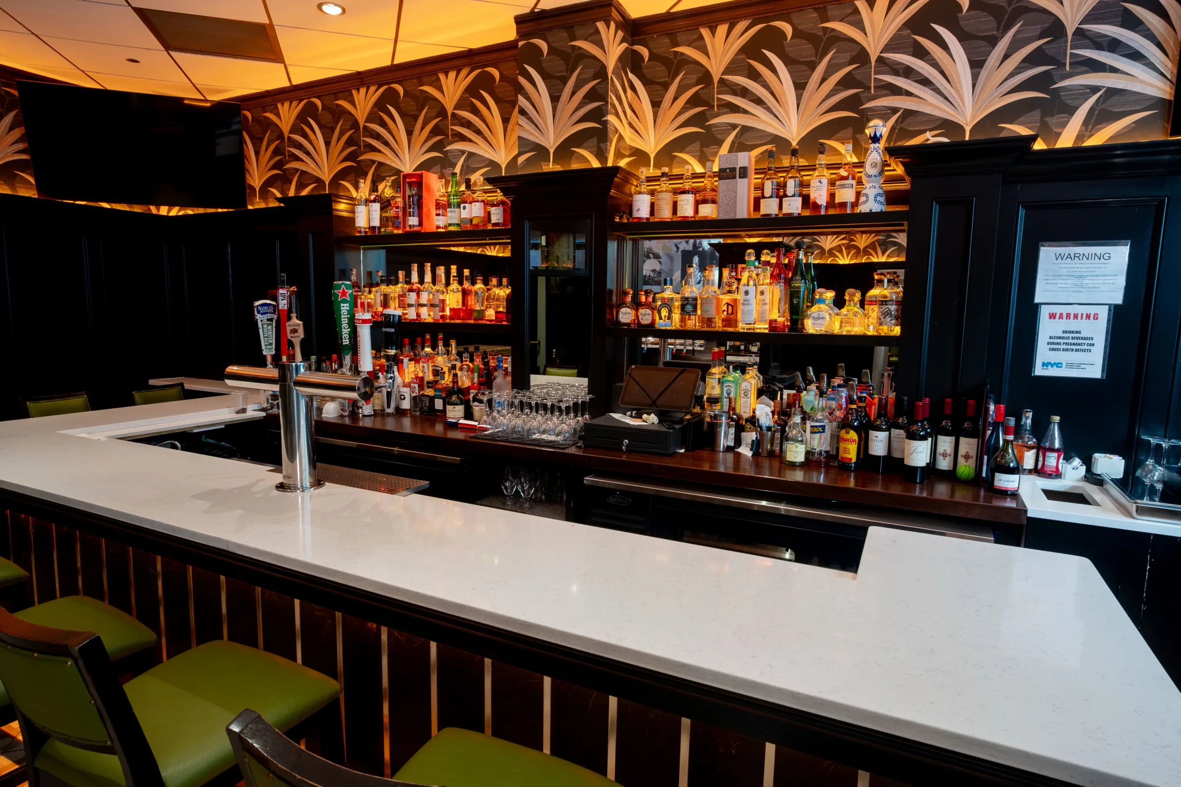 A white bar top curving in front of palm-leaf wallpaper and shelves of bottles, with green stools.