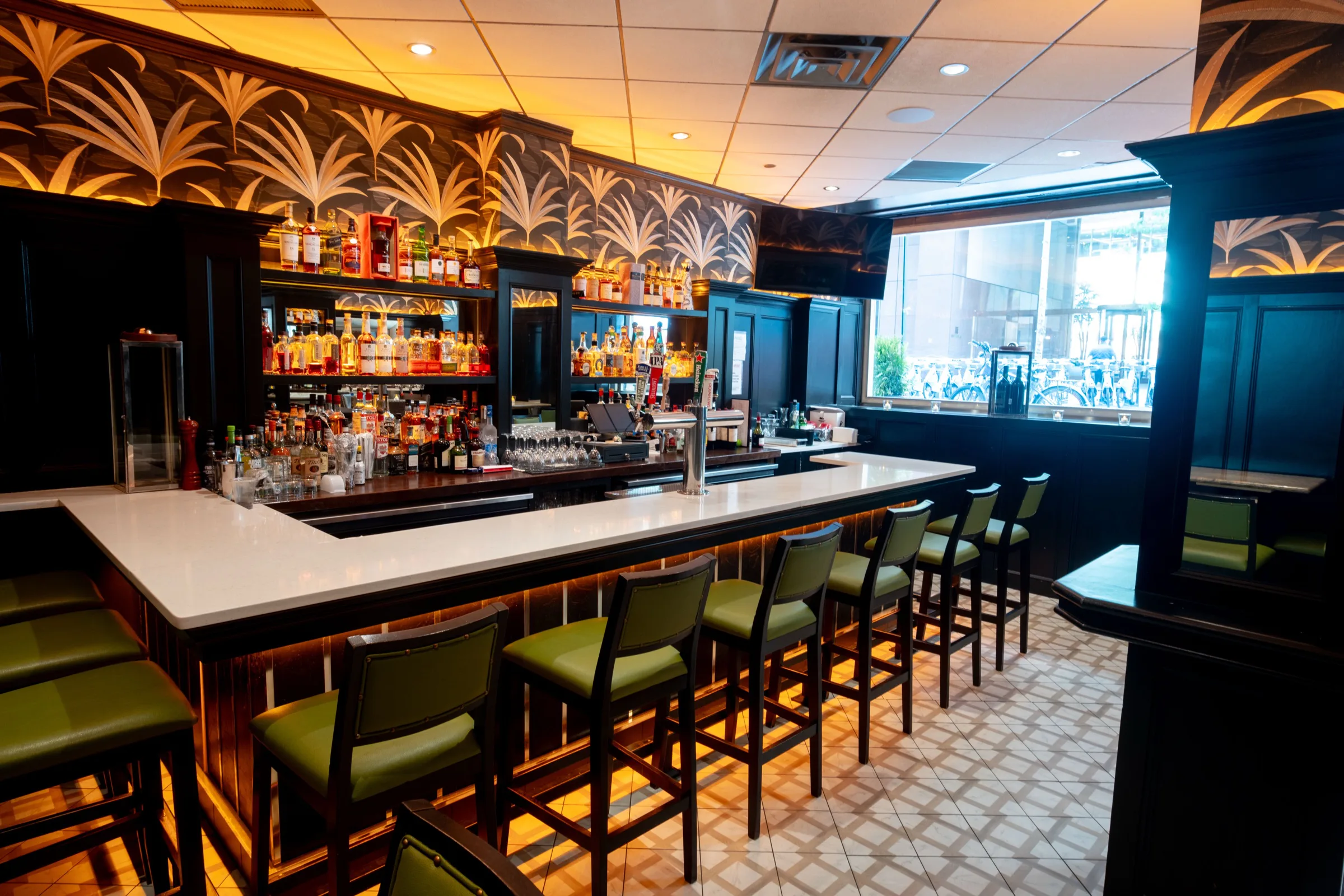 A long bar with green upholstered stools under palm-leaf wallpaper, with a black pillar and a window at the right.