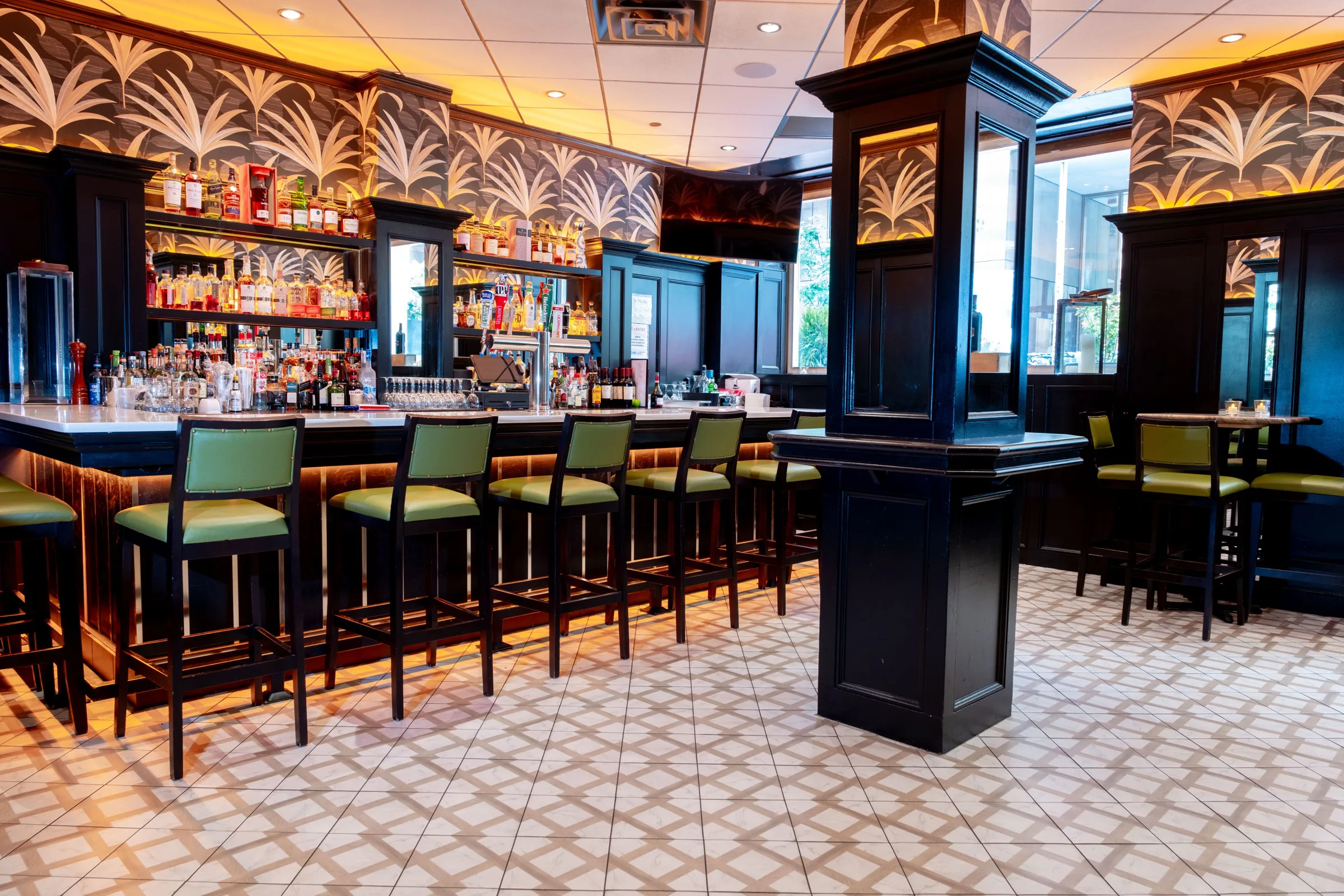 A bar with green stools and a black pillar on a patterned tile floor, under palm-leaf wallpaper.