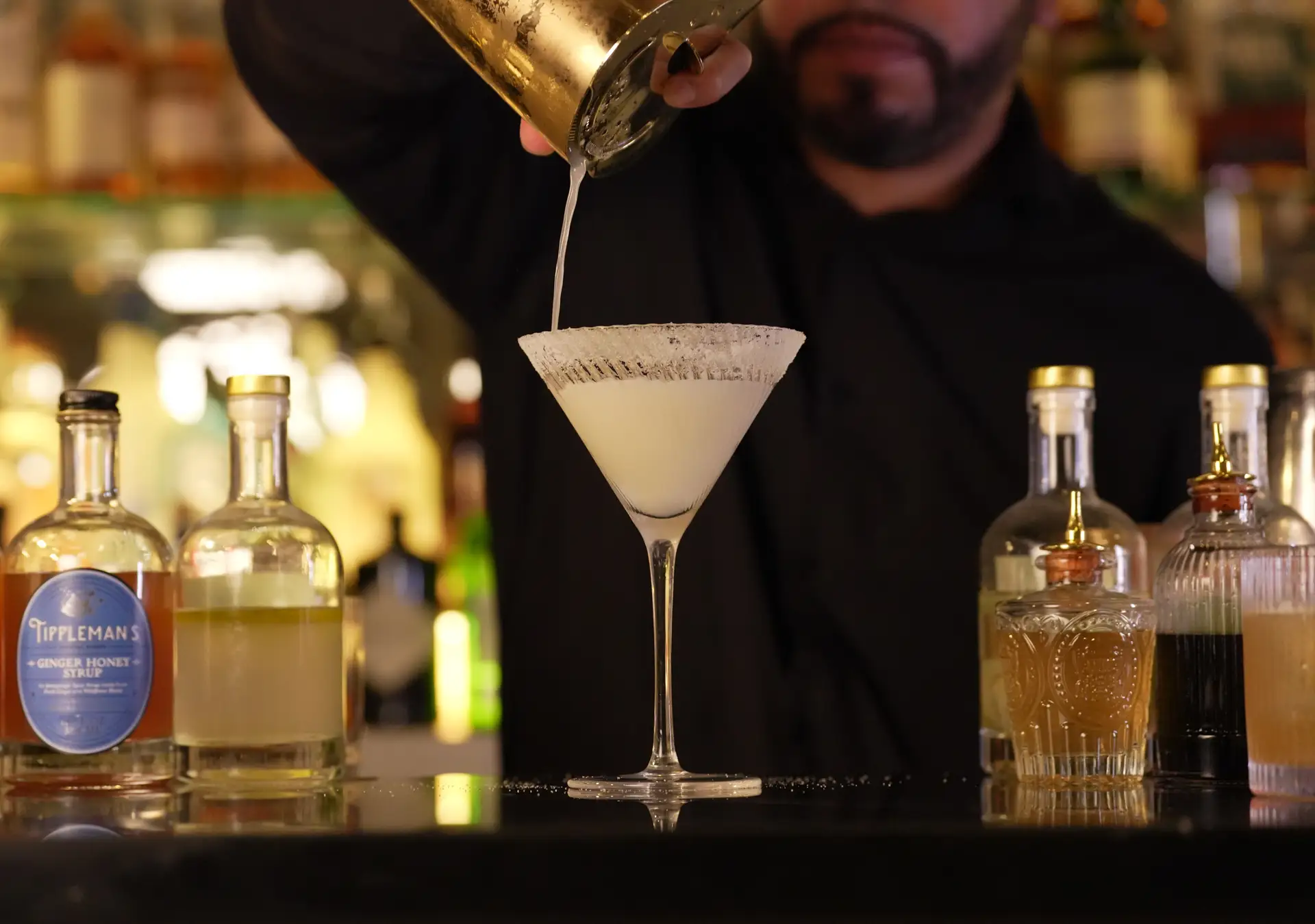 A bartender straining a pale cocktail into a martini glass on the bar